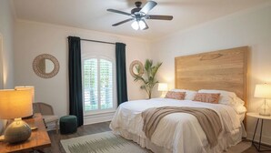 Ein Schlafzimmer mit einem großen Bett, zwei Nachttischlampen, einem Deckenventilator, einem Fenster mit Vorhängen, einem kleinen runden Spiegel und einem Holzkopfstück.