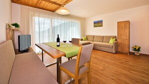 Ein Wohnzimmer mit Holzboden, einer Couch, einem Esstisch mit grüner Tischdecke, einem Fernseher und einem Schrank.