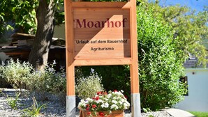 Ein hölzernes Schild mit der Aufschrift 'Moarhof' und 'Agriturismo', umgeben von Pflanzen und Kies.