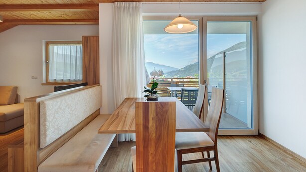 A wooden dining table with benches, a sofa, a TV, and a view of mountains through a large window. - Apartment 'Sonnenblume' with Mountain View, Shared Garden and Wi-Fi (Rasa)