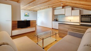 A modern living room with a glass coffee table, a sofa, a TV, and a kitchen area with cabinets and appliances. - Apartment 'Sonnenblume' with Mountain View, Shared Garden and Wi-Fi (Rasa)