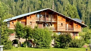 A multi-story log cabin with balconies, surrounded by lush greenery and a mountainous backdrop.