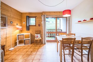 A wooden interior with a dining table, chairs, a lamp, and a view through a door.