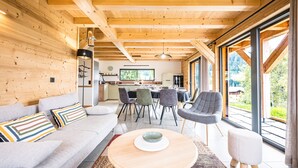 A modern interior with a wooden ceiling, a dining area with chairs, a sofa with patterned cushions, and large windows offering a view.
