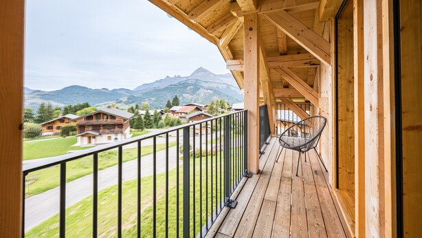 A wooden balcony with a metal railing, a hanging chair, and a view of houses and mountains.