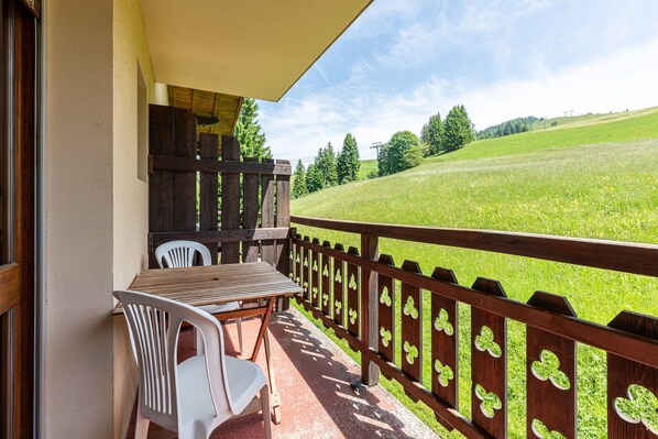 Een balkon met een houten tafel en stoelen, uitzicht op een groen veld, en een houten hek met klavertje vier uitsparingen.