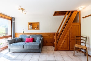 A room with a grey sofa, a wooden chair, a wooden staircase, and a small wooden shelf.