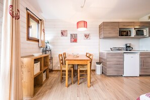 Een houten eettafel met stoelen, een kitchenette met een magnetron en een koffiezetapparaat, en een witte koelkast.