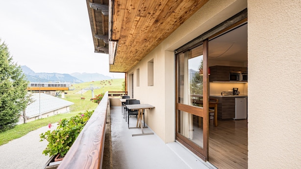 Un balcon doté d’un plafond en bois, un coin repas avec des tables et des chaises, et une cuisine visible à travers une porte vitrée.