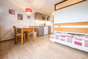 Een knus ingerichte kamer met een houten eettafel, stoelen, een bed met quilt, een kitchenette met magnetron, en een rode hanglamp.