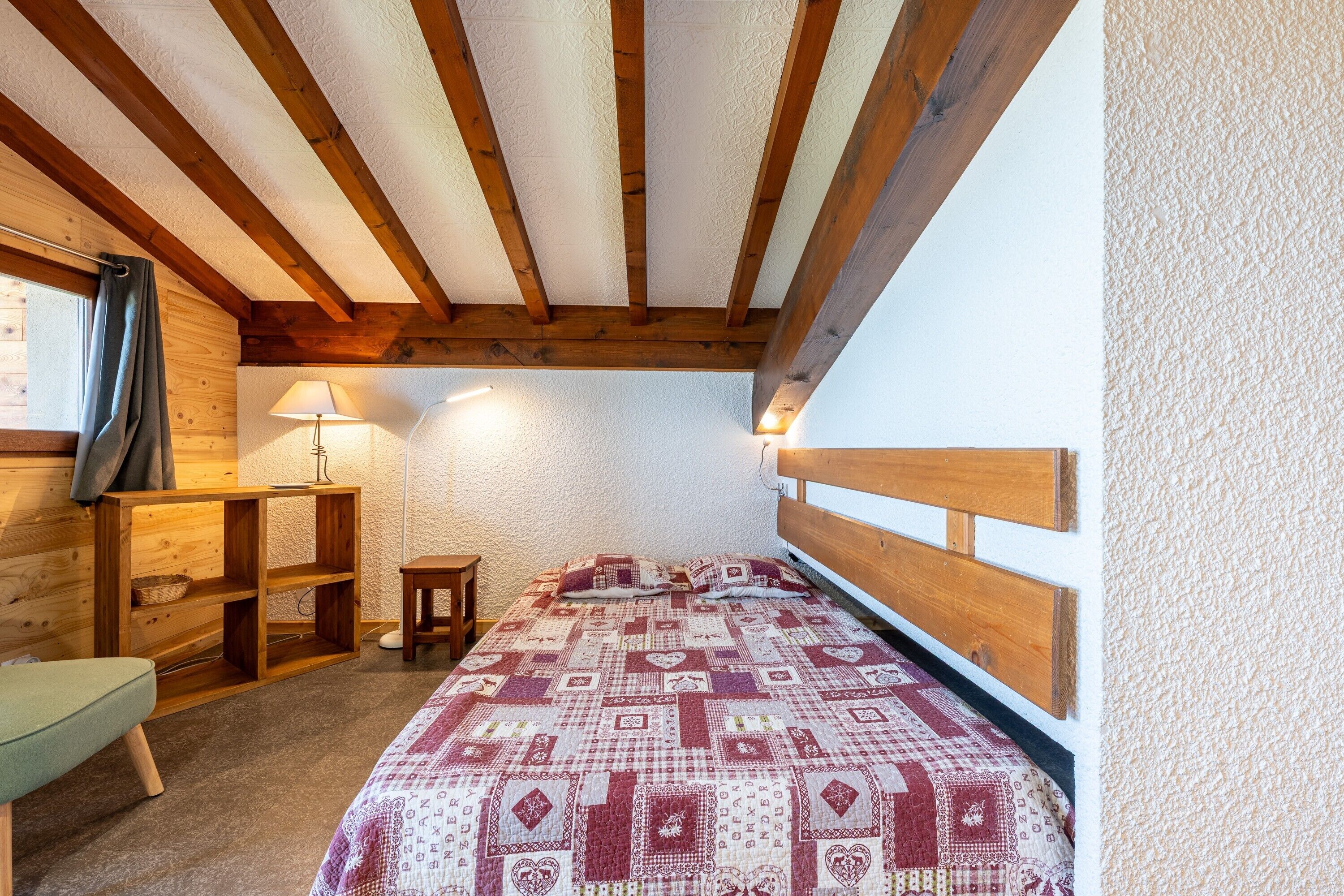 Une chambre avec un plafond en bois, un lit recouvert d’une couvre-lit à motifs, une table de chevet, une lampe et un bureau en bois doté d’étagères.