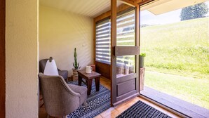 A room with a sliding glass door, a chair, a small table, a potted plant, and a view of a grassy field. - Studio Apartment '2 - L'Edelweiss' with Mountain View, Shared Garden and Wi-Fi (Cohennoz)