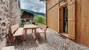 Uma mesa de madeira e bancos num pátio de calçada, com uma porta de madeira e uma parede de pedra.