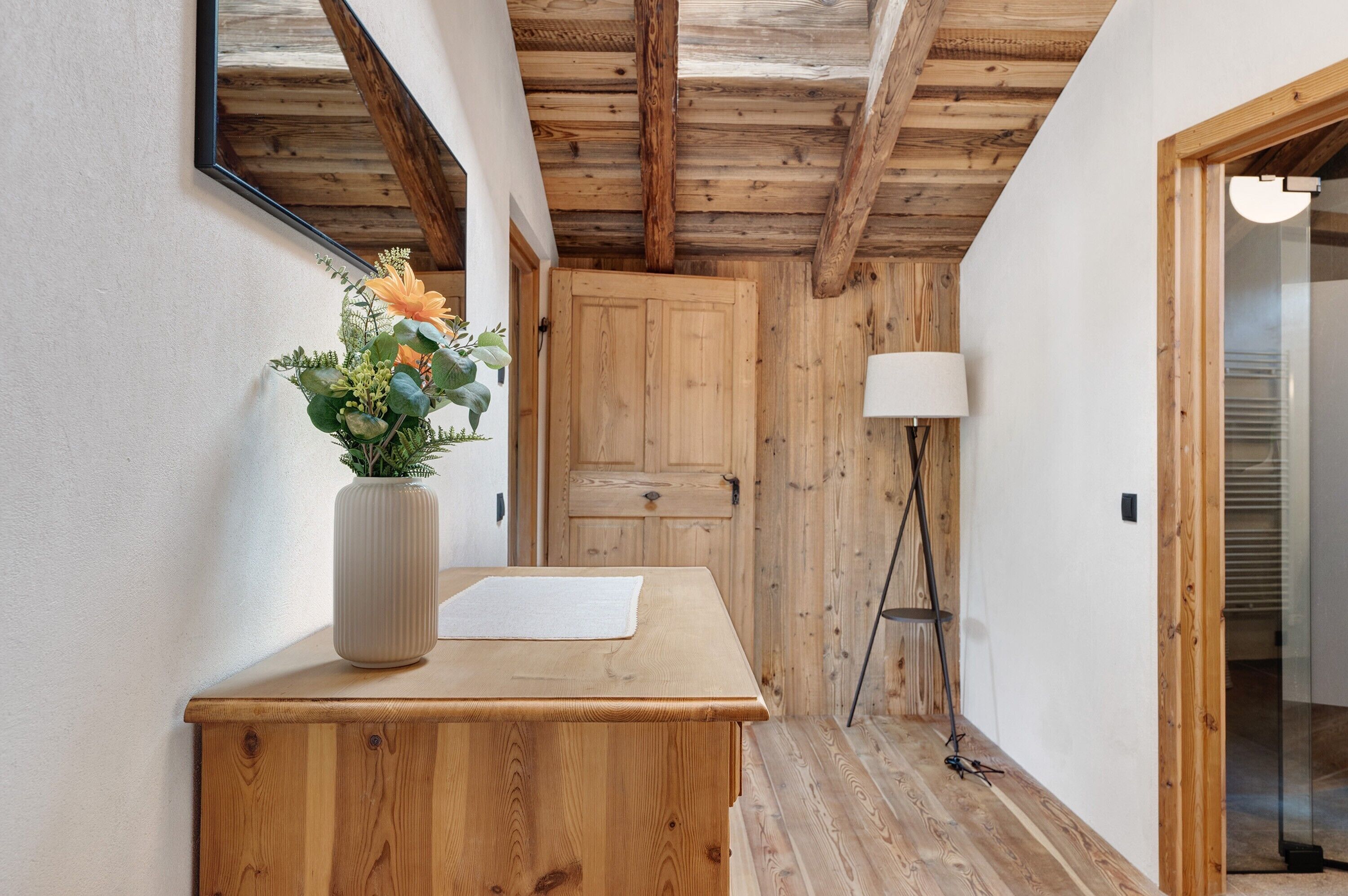 A wooden desk with a vase of flowers, a mirror, and a lamp in a hallway with wooden walls and a door.