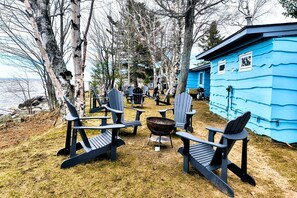 Une cabane bleue, entourĂ©e de chaises Adirondack disposĂ©es autour dâun foyer, le tout se dressant sur un fond de forĂȘt et avec un plan dâeau en arriĂšre-plan.