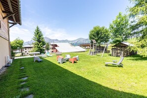 A grassy outdoor area with a wooden cabin, a play structure, and a view of mountains in the background.