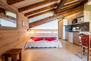 Un dormitorio acogedor con cabecera de madera, una mesita de noche pequeña y un almohadón rojo. Junto al dormitorio hay una cocina compacta con microondas, cafetera y un refrigerador pequeño.