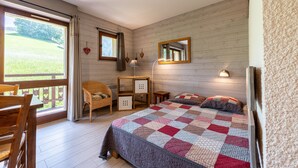 A bedroom with a wooden floor, a bed with a quilt, a chair, a small table, and a window with curtains. - Studio Apartment '10 - Bisanne' with Mountain View, Shared Garden and Wi-Fi (Cohennoz)