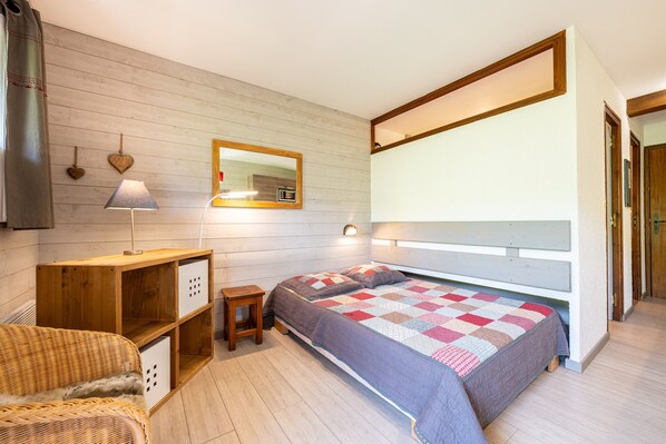 A bedroom with a wooden headboard, a bed with a quilt, a small wooden nightstand, a wicker chair, and a mirror on the wall.