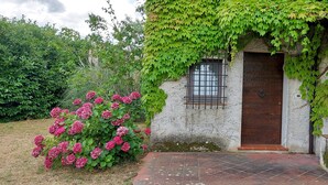 Un edificio rustico in pietra con una porta in legno e una piccola finestra, immerso nel verde e circondato da fiori rosa.