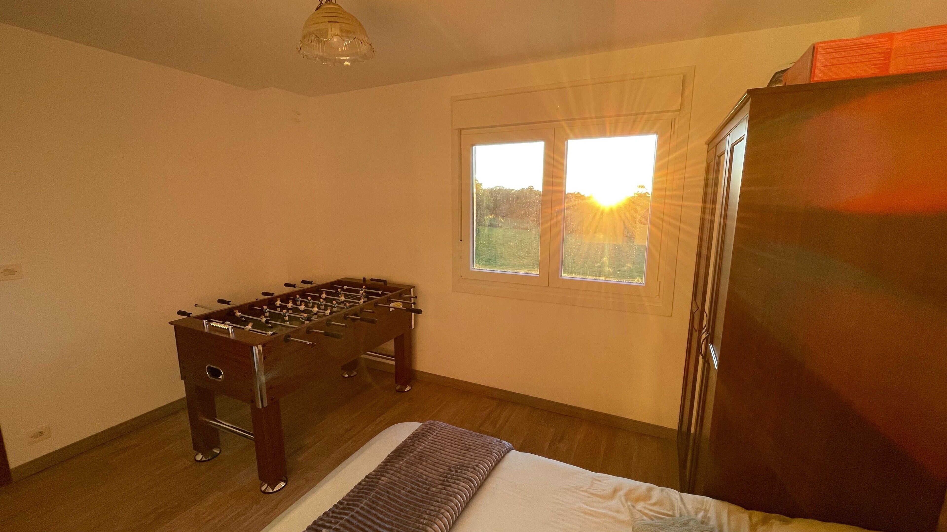 A room with a foosball table, a bed, a window with a view, and a refrigerator.