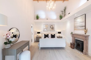 A modern bedroom with a large bed, bedside tables, a mirror, a fireplace, and artwork on the walls.