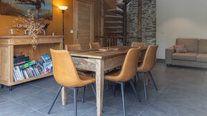 A dining area with a wooden table and chairs, a bookshelf with board games, a spiral staircase, and a sofa. - Chalet La Sentinelle – Alpine Elegance, Comfort, and Nature in the Heart of Tarentaise (Grand-Aigueblanche)