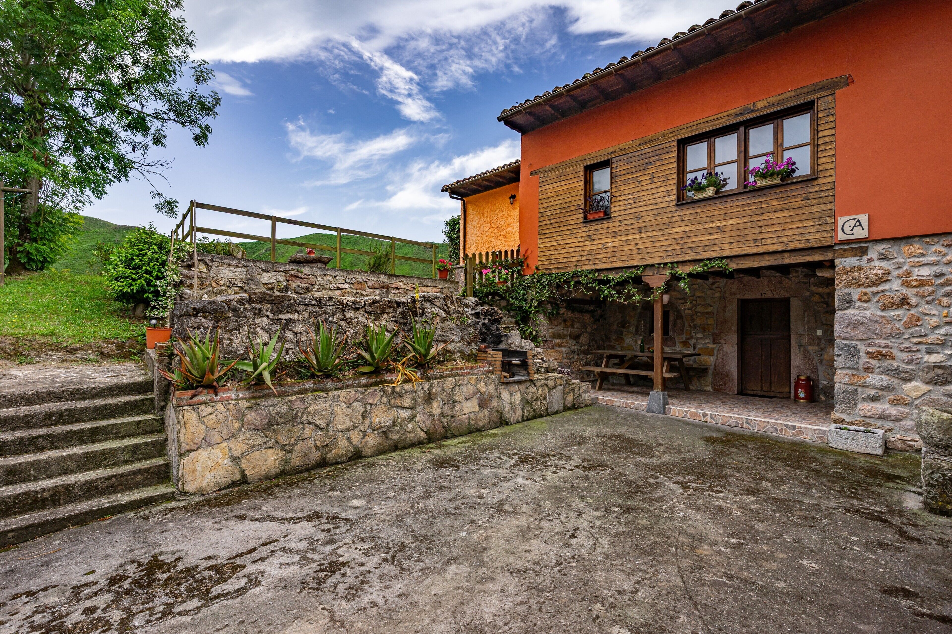 A rustic stone house with a wooden staircase, a stone wall with plants, and a wooden fence.