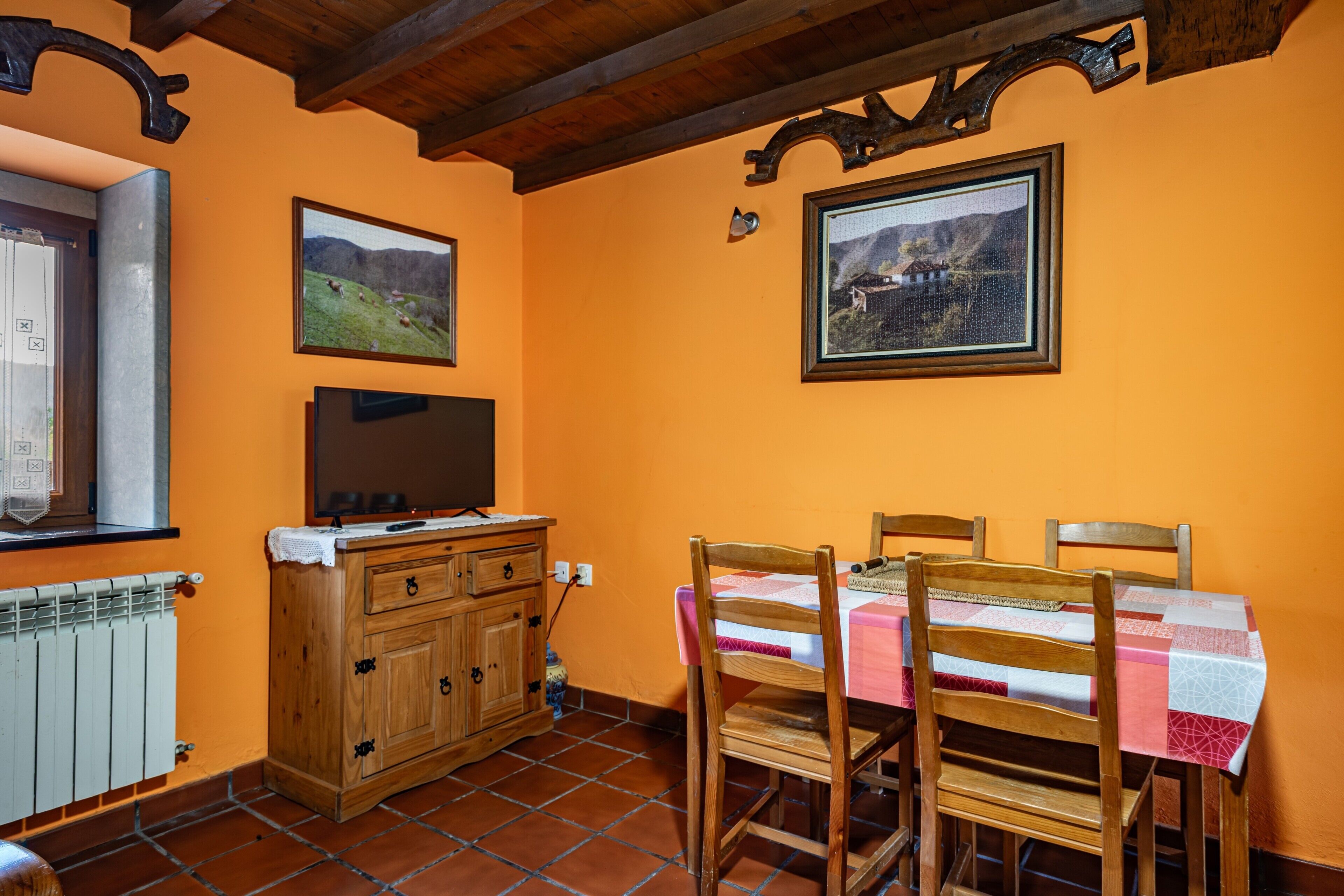 Un comedor con una mesa dispuesta para cuatro personas, sillas de madera, un aparador de madera con un televisor y dos cuadros en la pared.