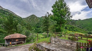 A rustic mountain cabin with a tiled roof, surrounded by lush greenery and a stone pathway leading to a wooden fence. - Casa Rural Ricao with Mountain Views, Private Terrace and WiFi (Cangas de onis)