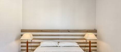 A minimalist bedroom with a wooden headboard, two bedside tables with lamps, a neatly made bed, and a white wall.