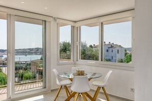 A modern room with a glass table, white chairs, and large windows offering a view of a body of water and buildings.