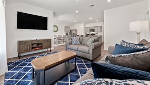 A modern living room with a sofa, coffee table, and TV. There is a kitchen in the background. - Brand-New 4BR Magnolia Retreat · Sleeps 8 · Fast WiFi · 30 Min to Woodlands (Magnolia)