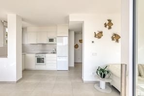 Een moderne keuken met witte kasten, een magnetron en een oven. Er staat een klein tafeltje met een plant en decoratieve wandkunst.