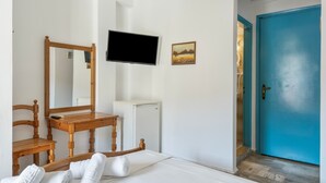 Un dormitorio con cama, una silla de madera, un escritorio, televisor, un refrigerador pequeño y una puerta azul.