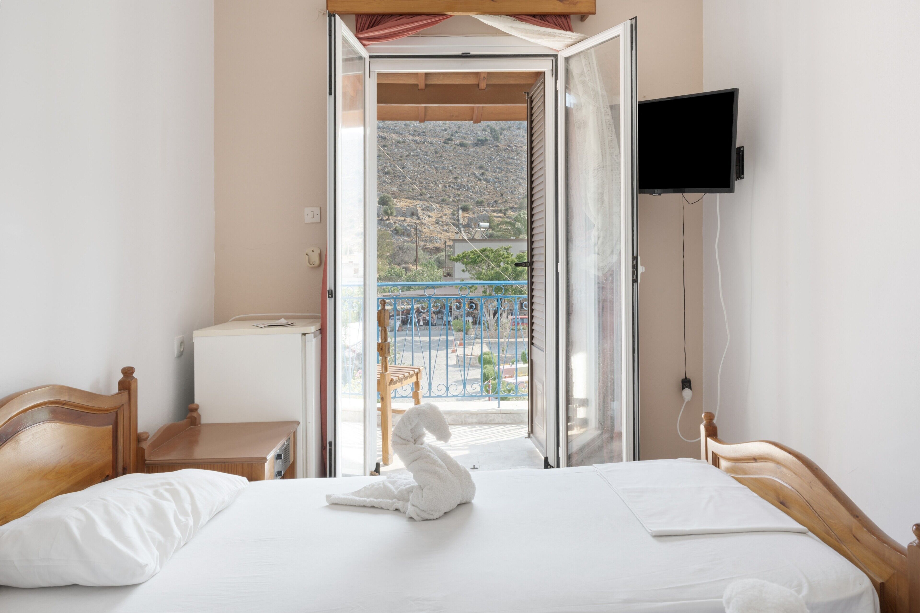 Habitación de hotel con cama, cabecera de madera, televisión montada en la pared y vistas a un balcón con barandilla.
