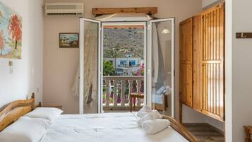 A bedroom with a bed, a balcony with a view of a mountain and a blue house, and a wooden wardrobe.