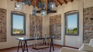 Une pièce avec une table en verre, deux fenêtres offrant une vue dégagée, des luminaires suspendus en métal et un mur en pierre.