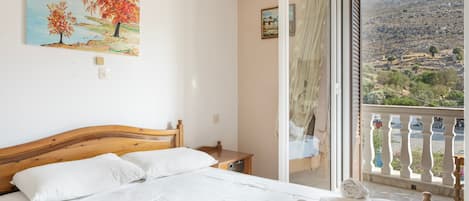 A bedroom with a wooden bed, white linens, a painting of trees, and a balcony with a view of a hilly landscape.