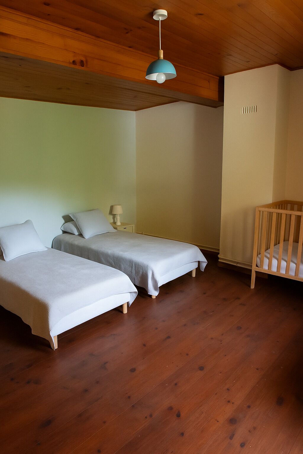 Une chambre avec deux lits, un berceau, un plancher en bois et un luminaire suspendu.