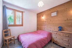 A bedroom with a bed, a wooden dresser, a chair, a window with curtains, and a wooden wall with a picture. - Chalet 'La Mouliniere 2' with Mountain View, Private Garden and Wi-Fi (Valloire)