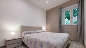 Une chambre à coucher comprenant un lit, une table de chevet, une lampe, une fenêtre avec des rideaux et une vue sur la verdure à l’extérieur.