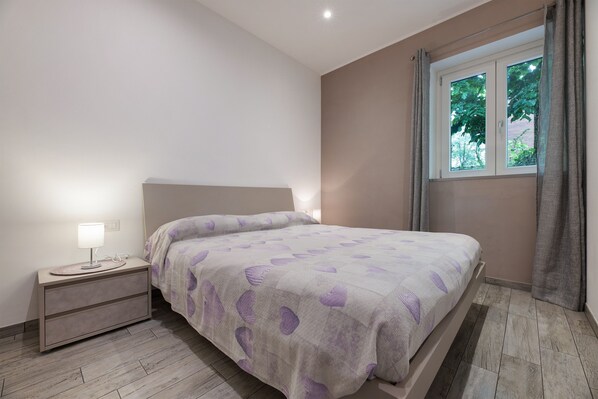 A bedroom with a bed, nightstand, lamp, window with curtains, and a view of greenery outside.