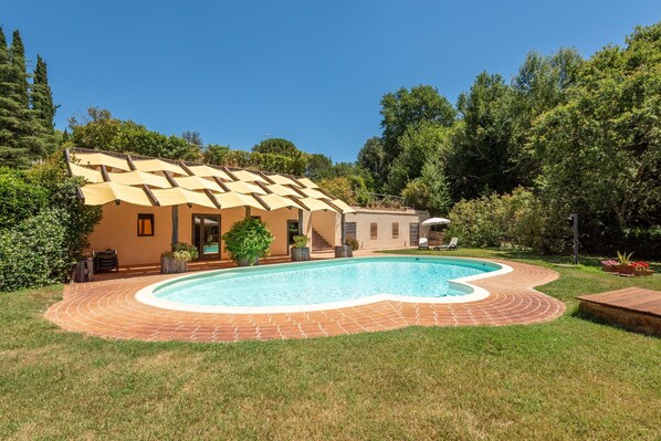 A swimming pool with a brick deck, a covered patio, and a house with a sloped roof.
