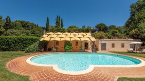 Ein Pool mit Backsteinrand, eine überdachte Terrasse und ein Gebäude mit beige gehauchter Fassade im Hintergrund.