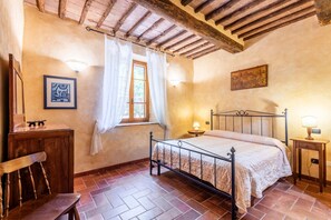 Une chambre à coucher comprenant un lit, une chaise en bois, une petite table, une fenêtre avec des rideaux et une lampe fixée au mur.