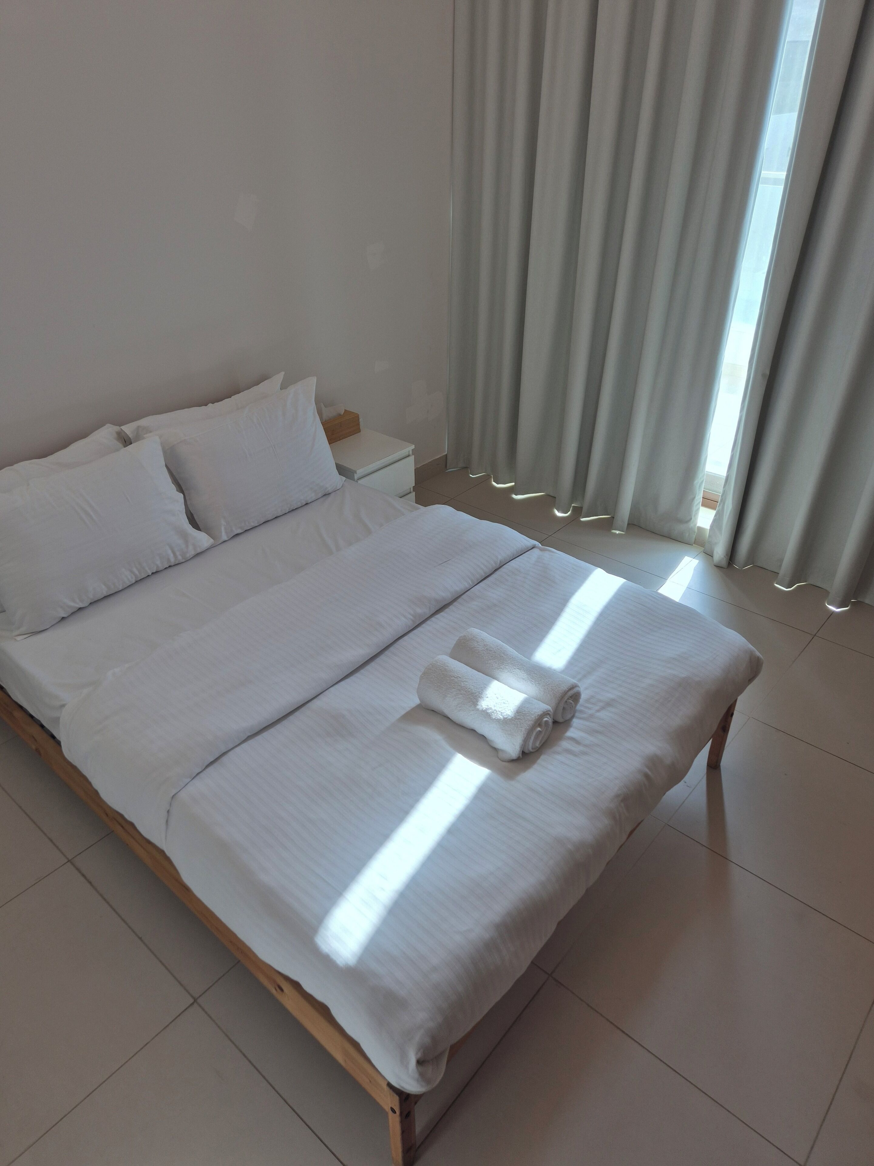 A neatly made bed with white linens and a rolled towel, a bedside table with a tissue box, and a window with curtains.