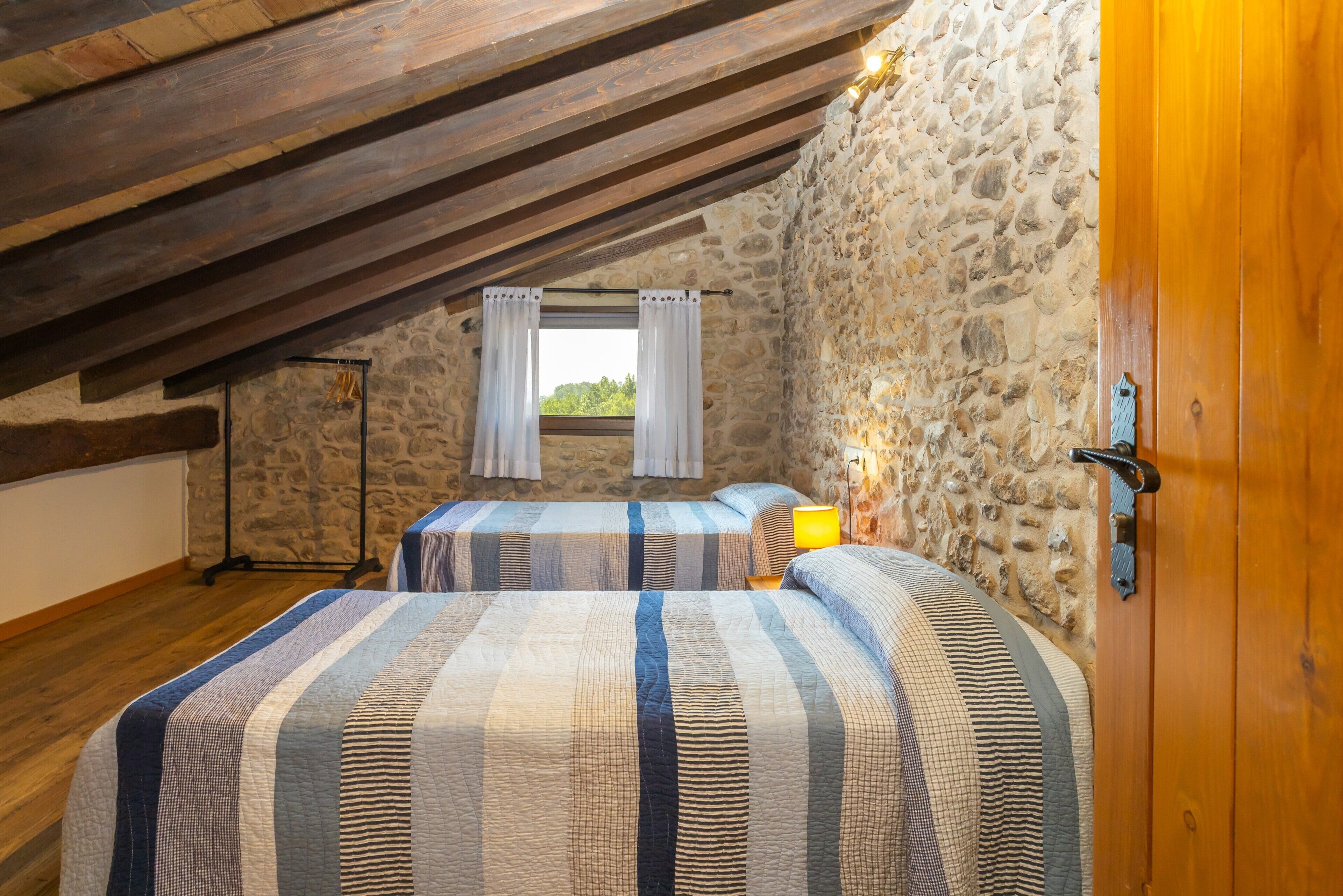 A bedroom with two beds, stone walls, wooden beams, a window with curtains, and a bedside lamp.