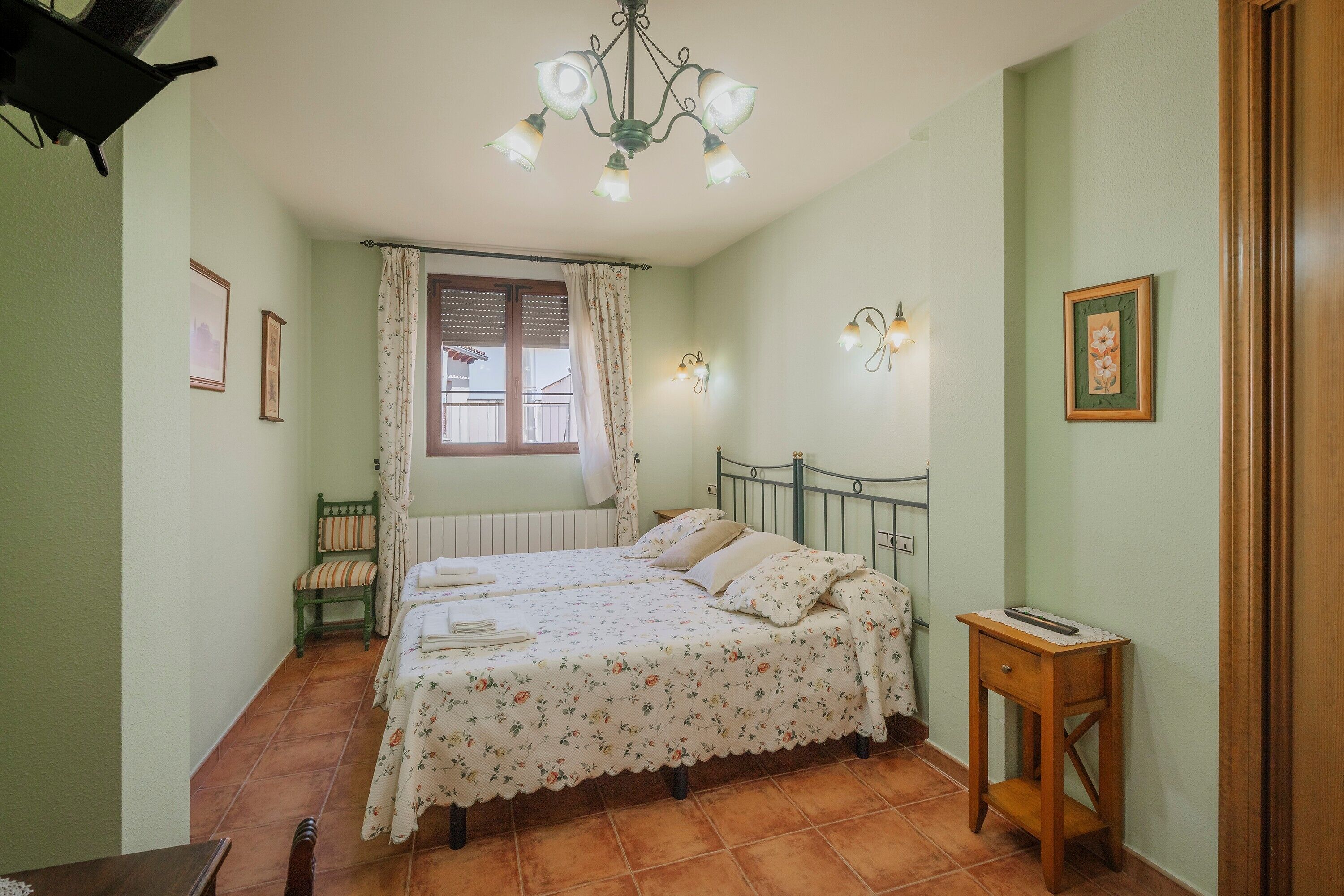 Un dormitorio con cama, una silla, una mesita, un ventanal con cortinas y un candelabro.
