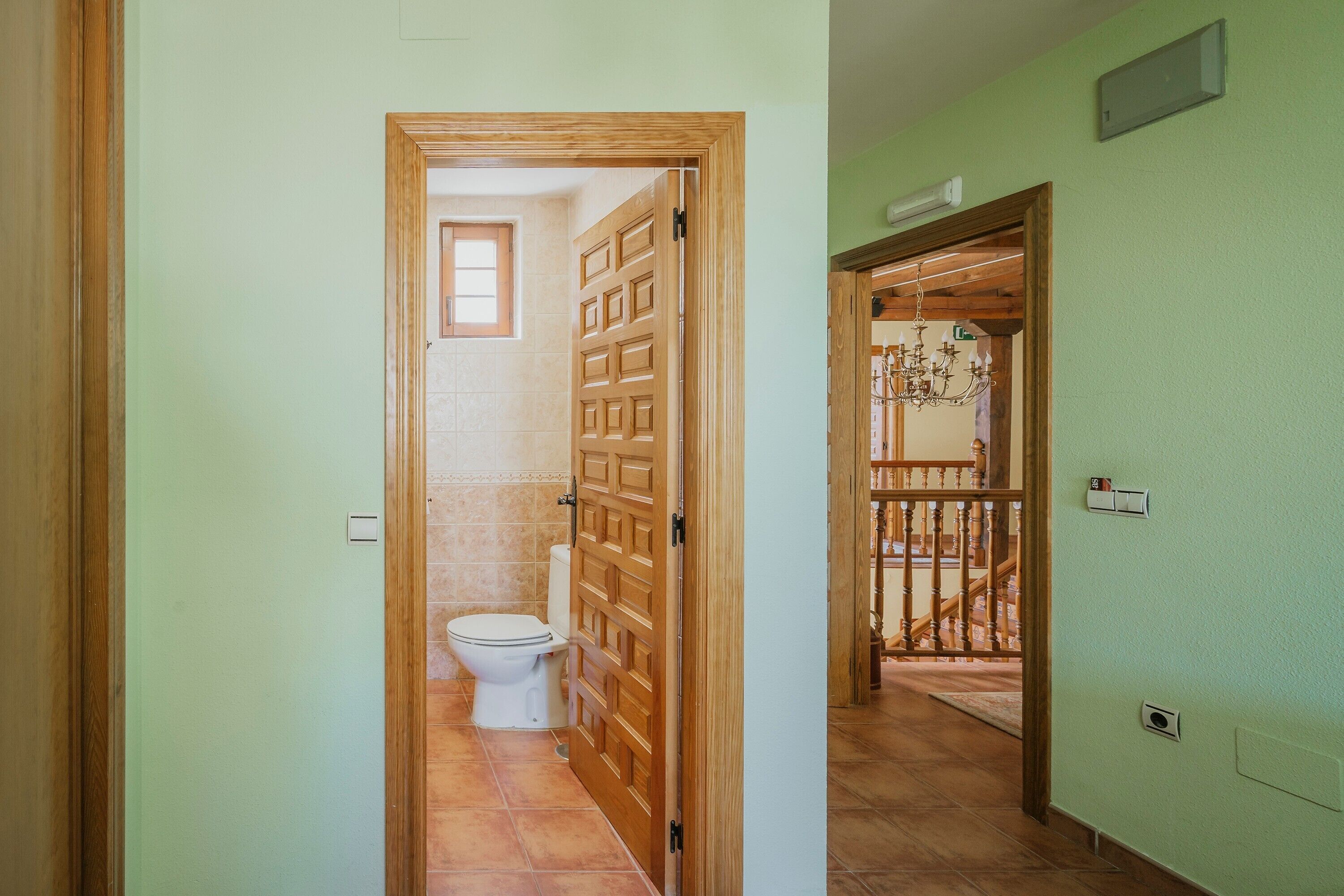 Un couloir avec une porte en bois donnant sur une salle de bain équipée de toilettes et dont le sol est carrelé.